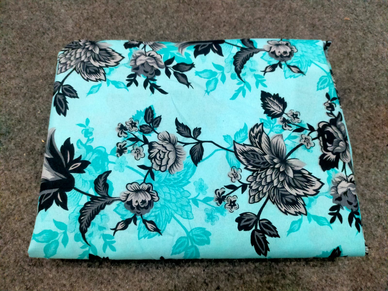 Floral-patterned fabric with black flowers on a turquoise background, folded on a brown surface.